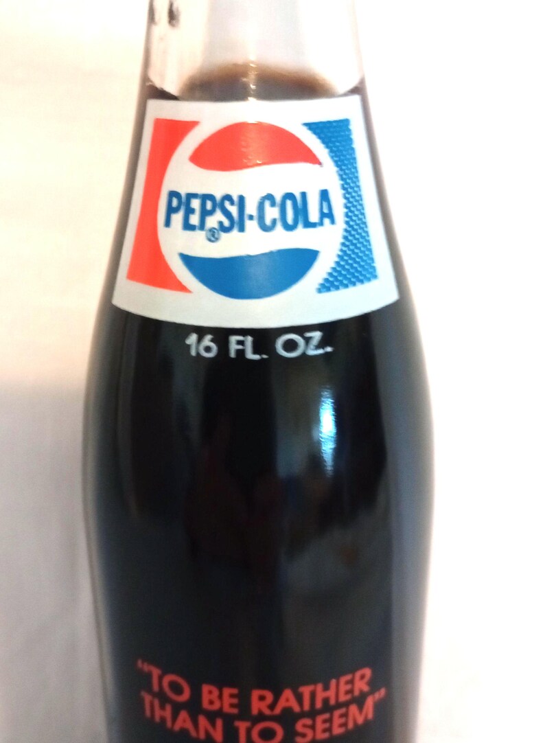 May include: A glass bottle of Pepsi-Cola with a red, white, and blue label. The bottle is 16 fluid ounces and has the quote "To be rather than to seem" printed on the bottom.