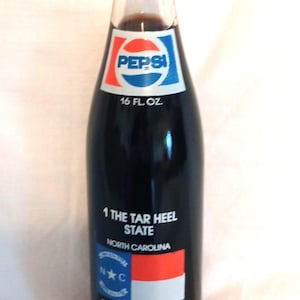 May include: A glass bottle of Pepsi-Cola with a label featuring the North Carolina state flag and the text "1 The Tar Heel State North Carolina". The label also includes the text "Flag adopted in 1885 the dates May 20, 1775 is the Mecklenburg Declaration and April 12, 1776 is the date North Carolina declared its independence."