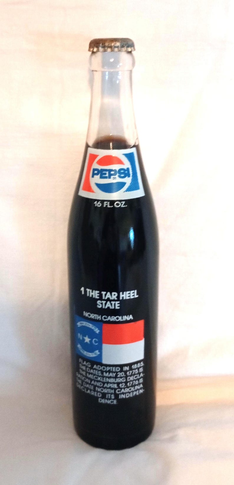May include: A glass bottle of Pepsi-Cola with a label featuring the North Carolina state flag and the text "1 The Tar Heel State North Carolina". The label also includes the text "Flag adopted in 1885 the dates May 20, 1775 is the Mecklenburg Declaration and April 12, 1776 is the date North Carolina declared its independence."