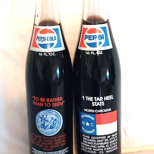 May include: Two 16-ounce Pepsi-Cola bottles with commemorative labels. The left bottle features the Great Seal of the State of North Carolina with the text "To be rather than to seem". The right bottle features the North Carolina state flag with the text "1 The Tar Heel State" and a description of the flag's history.