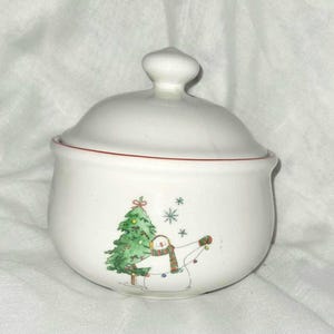 May include: White ceramic sugar bowl with a lid, featuring a Christmas tree and snowman design. The bowl has a red trim around the rim and a small knob on the lid. The snowman is wearing a scarf and holding lights.