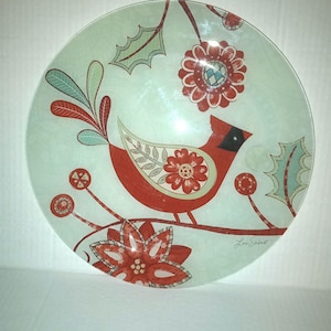 May include: A round glass serving dish with a red cardinal bird and floral design. The dish has a light blue background with green leaves and red flowers. The artist's signature "Lori Siebert" is in the bottom right corner.