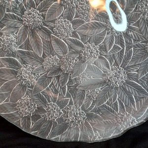 May include: A clear glass serving platter with a raised floral design. The design features leaves and flowers in a repeating pattern.