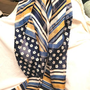 May include: A sheer scarf with a mix of patterns. The scarf features stripes in navy, white, and gold, along with sections of navy fabric adorned with small white flowers. The scarf is draped on a white surface.