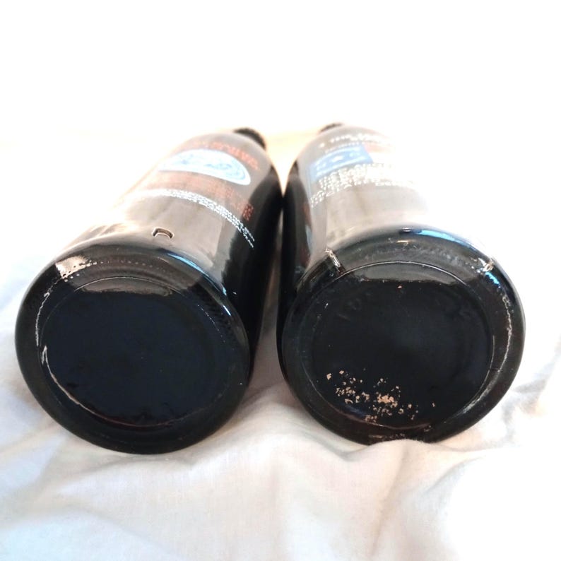May include: Two dark glass bottles with black caps. The bottles are empty and have a white label with black text.