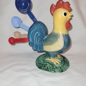 May include: A ceramic rooster-shaped measuring spoon holder. The rooster has a yellow body, blue tail, and red comb. It holds several colorful plastic measuring spoons. The base is green, resembling grass.