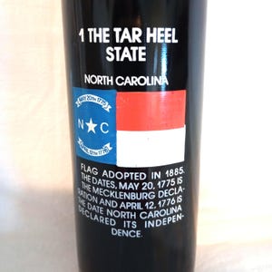 May include: A black bottle with a white label featuring the North Carolina state flag and text that reads "1 The Tar Heel State North Carolina Flag Adopted in 1885. The dates, May 20, 1775 is the Mecklenburg Declaration and April 12, 1776 is the date North Carolina declared its independence."