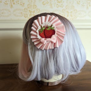 May include: A circular hair accessory featuring a pleated light pink ribbon base, a white lace trim, and an embroidered patch of two red strawberries with green leaves. A small light pink ribbon bow is attached to the bottom of the strawberry patch.