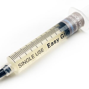 May include: A clear, single-use syringe filled with a pale liquid. The syringe has black measurement markings and the text "SINGLE USE" and "Easy Glide" printed on the side. A label reads "DRAGON OYS LOTUS EUNOS".