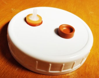 May include: A white plastic lid with two brown and orange bottle stoppers. One stopper has a white tube sticking out of it.