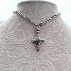 May include: A silver-toned necklace featuring a pendant in the shape of a four-pointed star with rounded edges and small spheres at each point. The pendant hangs from a delicate chain. The necklace is displayed on a mannequin.