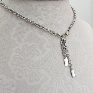 May include: A silver-toned metal chain necklace featuring a lobster claw clasp closure. The necklace includes two short chain segments hanging from the clasp, each finished with a small, smooth, rectangular metal tag at the end of the chain.