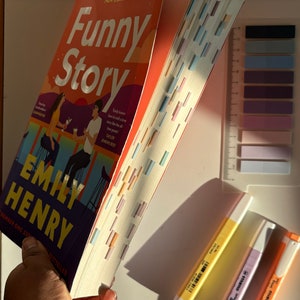 May include: A paperback book titled "Funny Story" by Emily Henry. The book is open and the pages are fanned out. The book cover features a couple sitting at a bar with a sunset in the background. There are also three highlighters in yellow, orange, and pink.