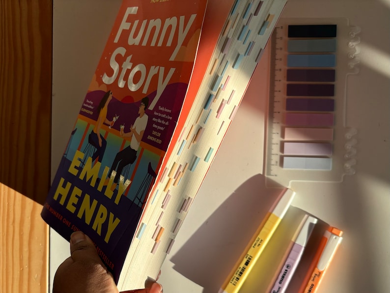 May include: A paperback book titled "Funny Story" by Emily Henry. The book is open and the pages are fanned out. The book cover features a couple sitting at a bar with a sunset in the background. There are also three highlighters in yellow, orange, and pink.