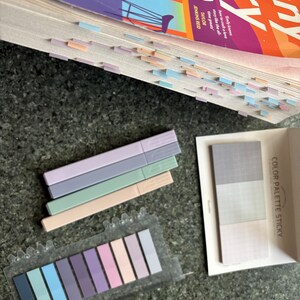 May include: A set of pastel colored sticky notes in a clear plastic dispenser. The notes are arranged in a rainbow pattern, with the darkest shade of blue on the left and the lightest shade of pink on the right. The notes are labeled "COLOR PALETTE STICKY".