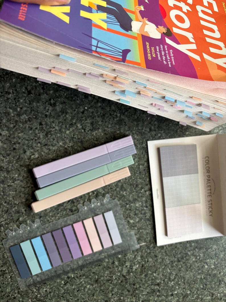 May include: A set of pastel colored sticky notes in a clear plastic dispenser. The notes are arranged in a rainbow pattern, with the darkest shade of blue on the left and the lightest shade of pink on the right. The notes are labeled "COLOR PALETTE STICKY".