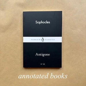 May include: A black book titled "Antigone" by Sophocles, part of the Penguin Classics series. The book features a white band with the Penguin logo. The text "annotated books" is at the bottom.