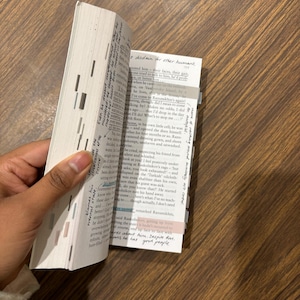 May include: An open book with highlighted text and sticky notes. The pages are white with black text. The book is held open by a hand, revealing the text and annotations. The book is on a wooden surface.