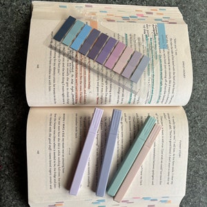 May include: A close-up of five pastel highlighters on a page of a book. The highlighters are in shades of lavender, light blue, and pink. The book is open to a page with text about a character named "Emily Henry".