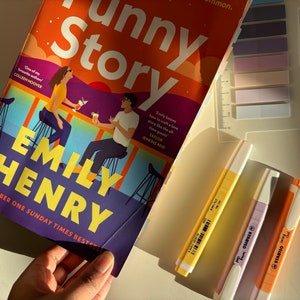 May include: A paperback book titled "Funny Story" by Emily Henry. The cover features an illustration of two people sitting at a bar with a sunset in the background. The book is a Sunday Times bestseller.