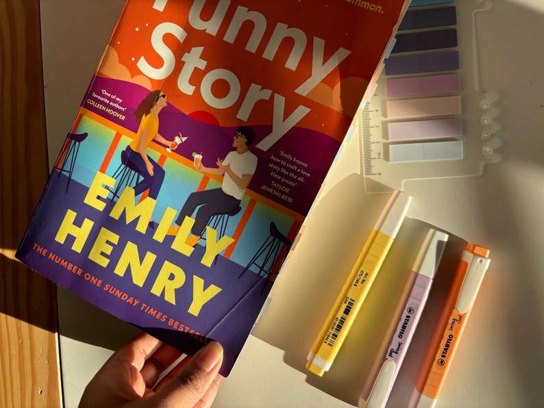 May include: A paperback book titled "Funny Story" by Emily Henry. The cover features an illustration of two people sitting at a bar with a sunset in the background. The book is a Sunday Times bestseller.