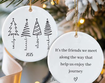 May include: White ceramic Christmas ornament with a gold string. One side features four stylized Christmas trees with names and the year 2025. The other side has the text: "It's the friends we meet along the way that help us enjoy the journey."