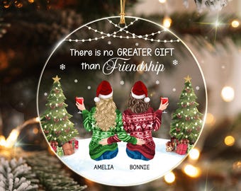 May include: A round clear acrylic Christmas ornament featuring two people from behind, wearing Santa hats and festive sweaters, holding red glasses. The text reads: There is no GREATER GIFT than Friendship, with the names AMELIA and BONNIE below.