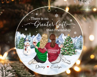 May include: A round, clear acrylic Christmas ornament featuring two people sitting in the snow, one in a green sweater and one in a red patterned sweater. The text reads, There is no Greater Gift than Friendship, with the names Daisy and Evelyn.