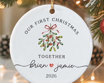 Può includere: Una decorazione natalizia rotonda in ceramica bianca con un'illustrazione di vischio e un fiocco rosso. Il testo recita Our First Christmas Together, i nomi brian e jamie separati da un cuore rosso, e l'anno 2026. Appesa a un cordino dorato.
