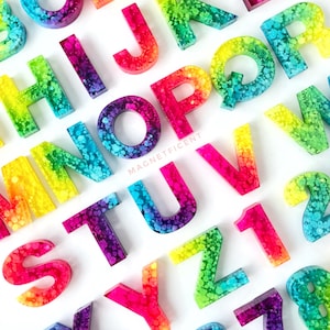 May include: Rainbow alphabet and number letter tiles made of resin with a glittery finish. The letters are in a rainbow gradient with a white background.