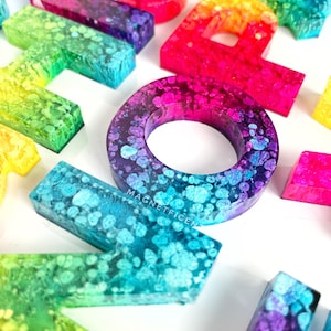 May include: A set of colorful resin alphabet letters with a rainbow gradient and white speckles. The letters are arranged in a random order and are all uppercase. The letters are made of resin and have a glossy finish.
