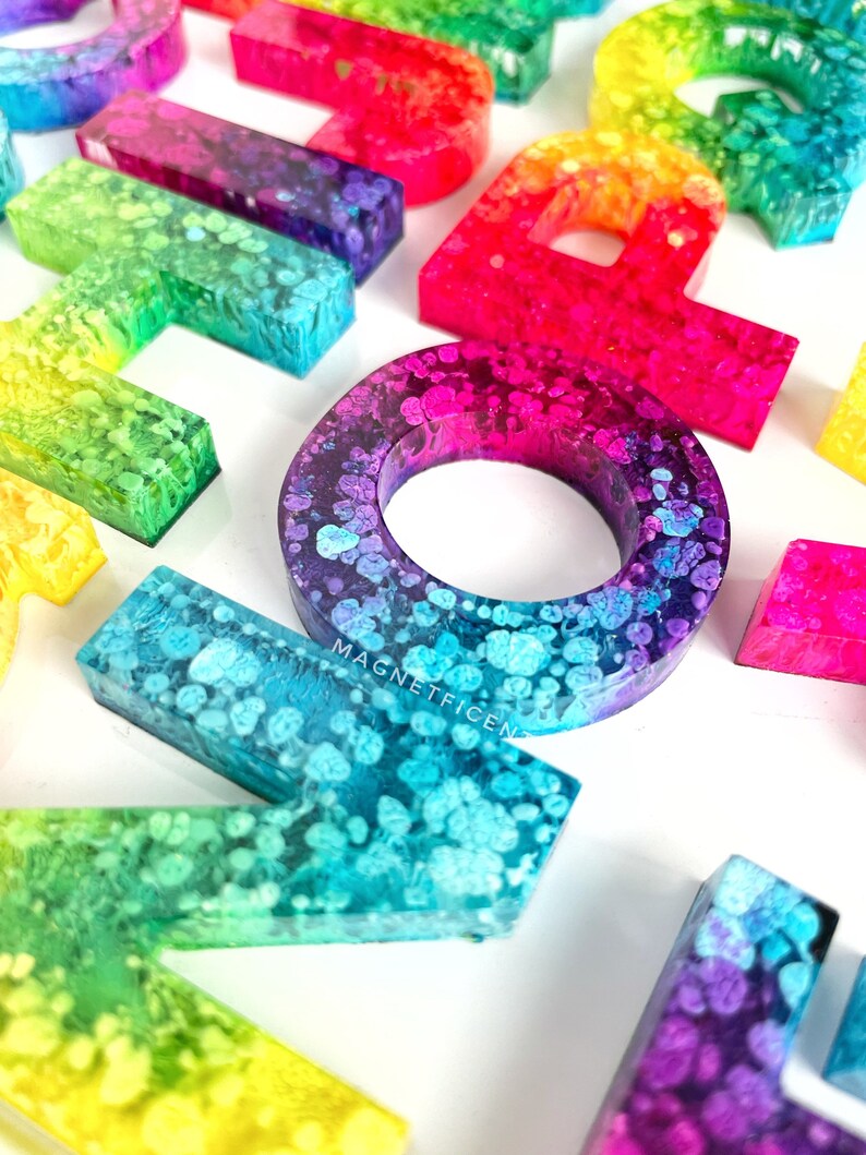 May include: A set of colorful resin alphabet letters with a rainbow gradient and white speckles. The letters are arranged in a random order and are all uppercase. The letters are made of resin and have a glossy finish.