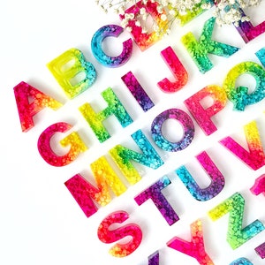 May include: Rainbow alphabet letters made of resin with a speckled, glittery finish. Each letter is a different color of the rainbow.