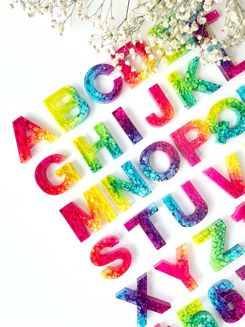 May include: Rainbow alphabet letters made of resin with a speckled, glittery finish. Each letter is a different color of the rainbow.