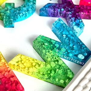 May include: A set of colorful resin alphabet letters with a rainbow gradient and white glitter. The letters are arranged in a random order on a white surface. The letters are all uppercase and are about 2 inches tall. The letters are made of resin and have a glossy finish. The letters are perfect for adding a touch of whimsy to any space.