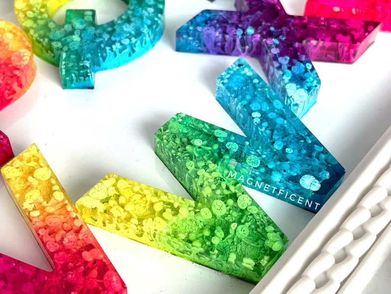 May include: A set of colorful resin alphabet letters with a rainbow gradient and white glitter. The letters are arranged in a random order on a white surface. The letters are all uppercase and are about 2 inches tall. The letters are made of resin and have a glossy finish. The letters are perfect for adding a touch of whimsy to any space.