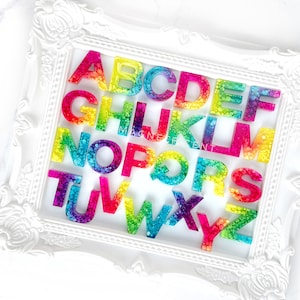 May include: A white frame with a rainbow alphabet made of resin. Each letter has a different color gradient and is filled with small, clear, sparkly dots.