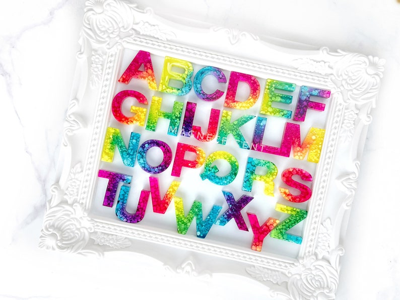 May include: A white frame with a rainbow alphabet made of resin. Each letter has a different color gradient and is filled with small, clear, sparkly dots.
