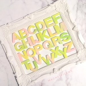 May include: A white frame with a decorative design features a colorful alphabet in a gradient of pink and green. The letters are arranged in alphabetical order and the frame has the word "MAGNETFICENT" printed on the bottom.