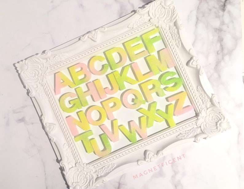 May include: A white frame with a decorative design features a colorful alphabet in a gradient of pink and green. The letters are arranged in alphabetical order and the frame has the word "MAGNETFICENT" printed on the bottom.