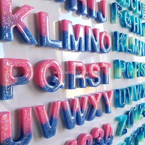 May include: A set of colorful, glittery, resin alphabet letters and numbers. The letters are two-toned, with pink on top and blue on the bottom. The numbers are also two-toned, with pink on top and blue on the bottom. The letters and numbers are arranged in alphabetical and numerical order.
