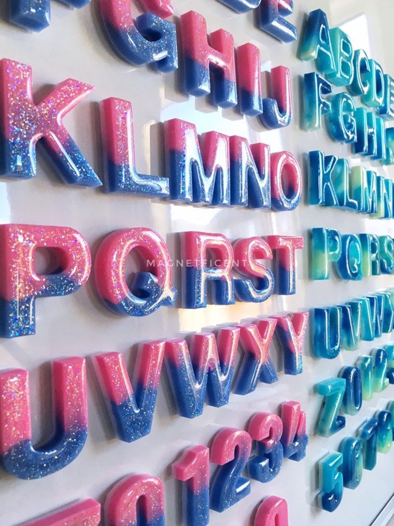 May include: A set of colorful, glittery, resin alphabet letters and numbers. The letters are two-toned, with pink on top and blue on the bottom. The numbers are also two-toned, with pink on top and blue on the bottom. The letters and numbers are arranged in alphabetical and numerical order.