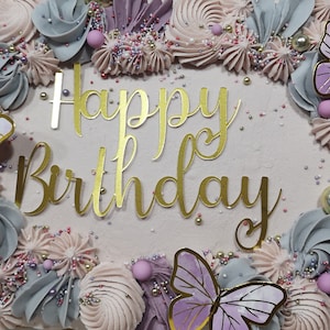 May include: A pink and purple frosted cake decorated with buttercream swirls, sprinkles, and gold glitter. The cake has a gold glitter 'Happy Birthday' message on top.