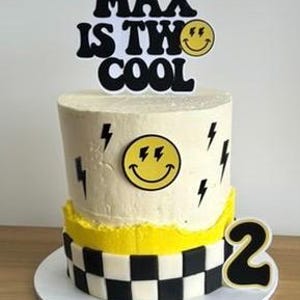 May include: A two-tiered birthday cake with white frosting, yellow accents, and a checkered bottom layer. The cake is decorated with black lightning bolts and a yellow smiley face. A cake topper reads "MAX IS TWO COOL".