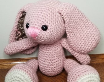 Könnte beinhalten: Ein handgehäkelter, rosa Hase mit langen Schlappohren. Der Hase hat schwarze Sicherheitsaugen, eine rosa Nase und weiße Akzente an den Füßen. Die Innenseite der Ohren ist mit einem braunen und cremefarbenen Blumenstoff versehen. Der Hase sitzt auf einer Holzoberfläche.
