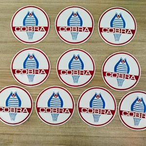 May include: Nine white round stickers with a red border and a blue cobra logo. The logo features a stylised cobra with a red tongue and the word "COBRA" in red letters.