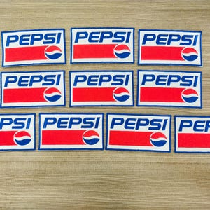 Könnte beinhalten: Zehn blaue und weiße rechteckige Patches mit dem Wort "PEPSI" in blauen Buchstaben und einem roten Streifen in der Mitte. Jeder Patch hat ein blaues und weißes Logo mit einem Kreis und einer Welle.