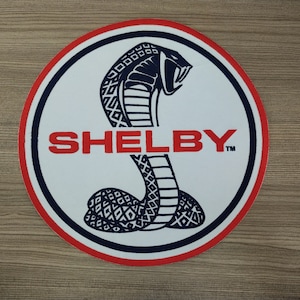 May include: A white circle with a red border and a blue and black cobra with the word "SHELBY" in red letters.