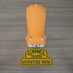 May include: A yellow and green patch with the text "Camel Trophy Adventure Wear" on a wooden surface. The patch is rectangular with rounded corners and a green border. The text is in a bold, white font.