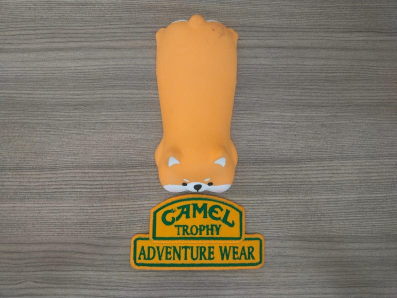 May include: A yellow and green patch with the text "Camel Trophy Adventure Wear" on a wooden surface. The patch is rectangular with rounded corners and a green border. The text is in a bold, white font.